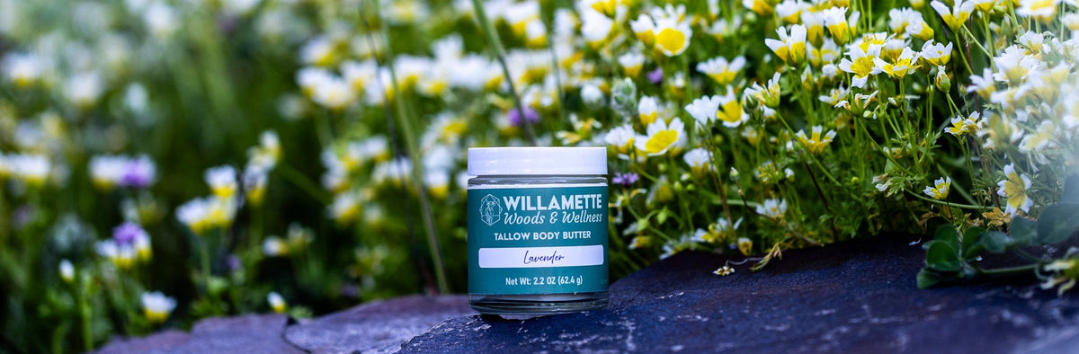 A small jar of Willamette Natural & Beautiful Coconut Body Butter sits on a dark stone surface in a field of white and yellow wildflowers.